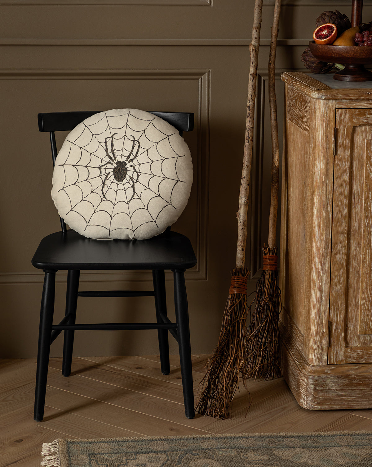 The Spider Pillow, featuring an embroidered spider design, rests on a black wooden chair while two rustic brooms lean against a cabinet—creating a cozy autumn or Halloween accent ideal for seasonal décor, by Vendixs