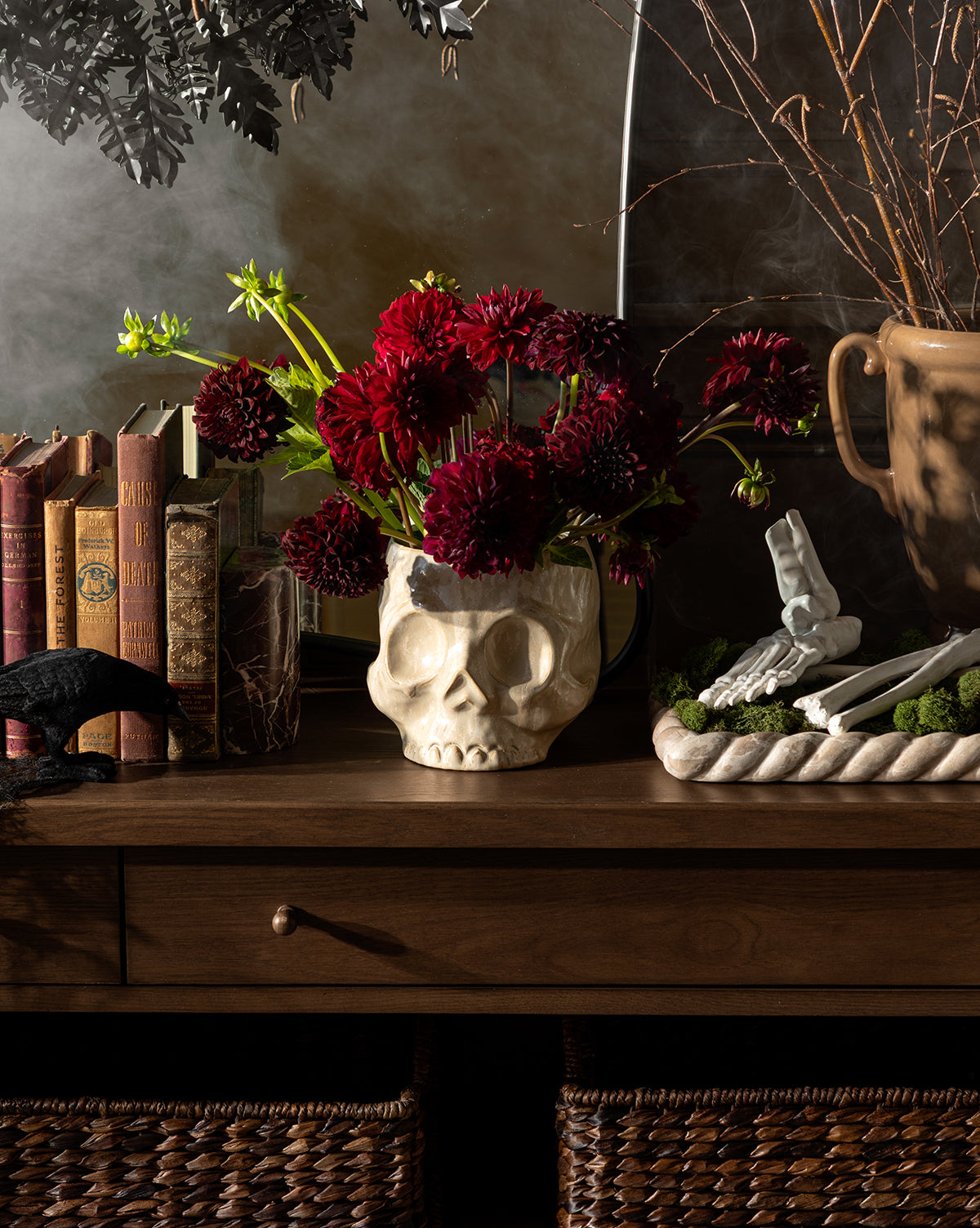 The Haunting Skull Vase, filled with dark red flowers, sits on a wooden table beside old books, a skeleton hand centerpiece, and a large urn—making it an unforgettable statement piece for dramatic Halloween décor, by Vendixs