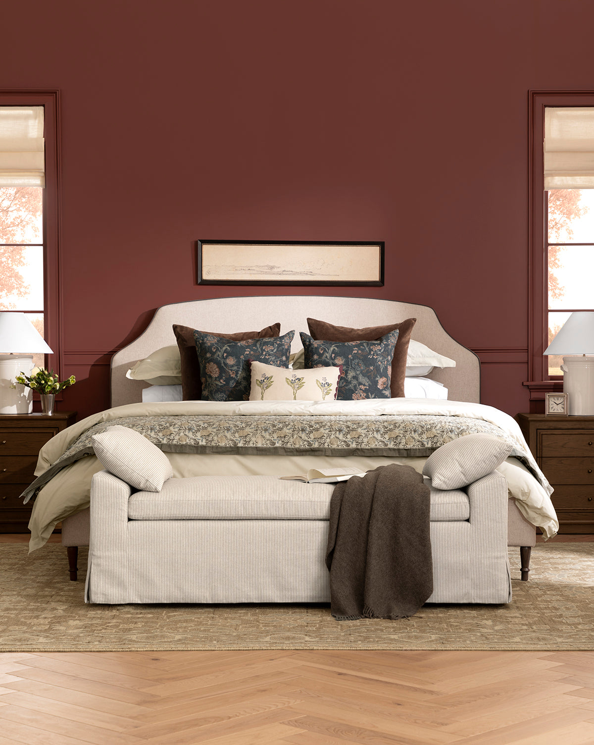 A cozy bedroom featuring a Willoughby Bed with linen upholstery, decorative pillows, and an English Oak bench at the foot. Wooden nightstands with lamps flank the bed, art hangs above, and deep red walls complete the inviting space, by Vendixs