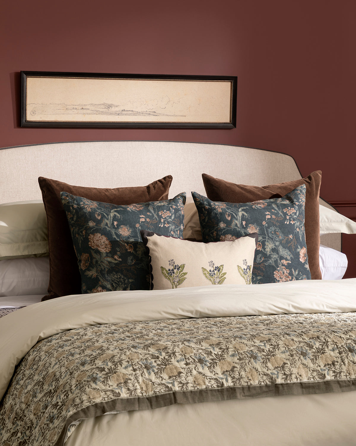 A neatly made bed features floral-patterned pillows and comforter, brown Bellevue Velvet Pillow Covers, a cream headboard, and a narrow framed artwork hanging on the mauve wall above the bed, by Vendixs