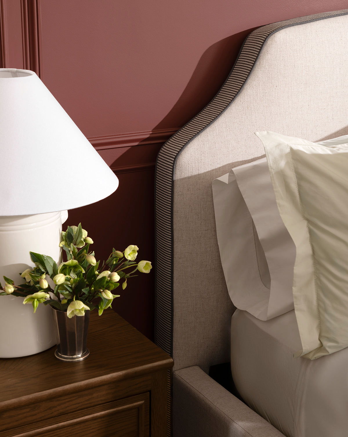 A close-up of a bedroom showcases the Willoughby Bed with linen upholstery, accented by white and light green pillows, a wooden nightstand, a white lamp, and a small flower vase against a mauve wall, by Vendixs