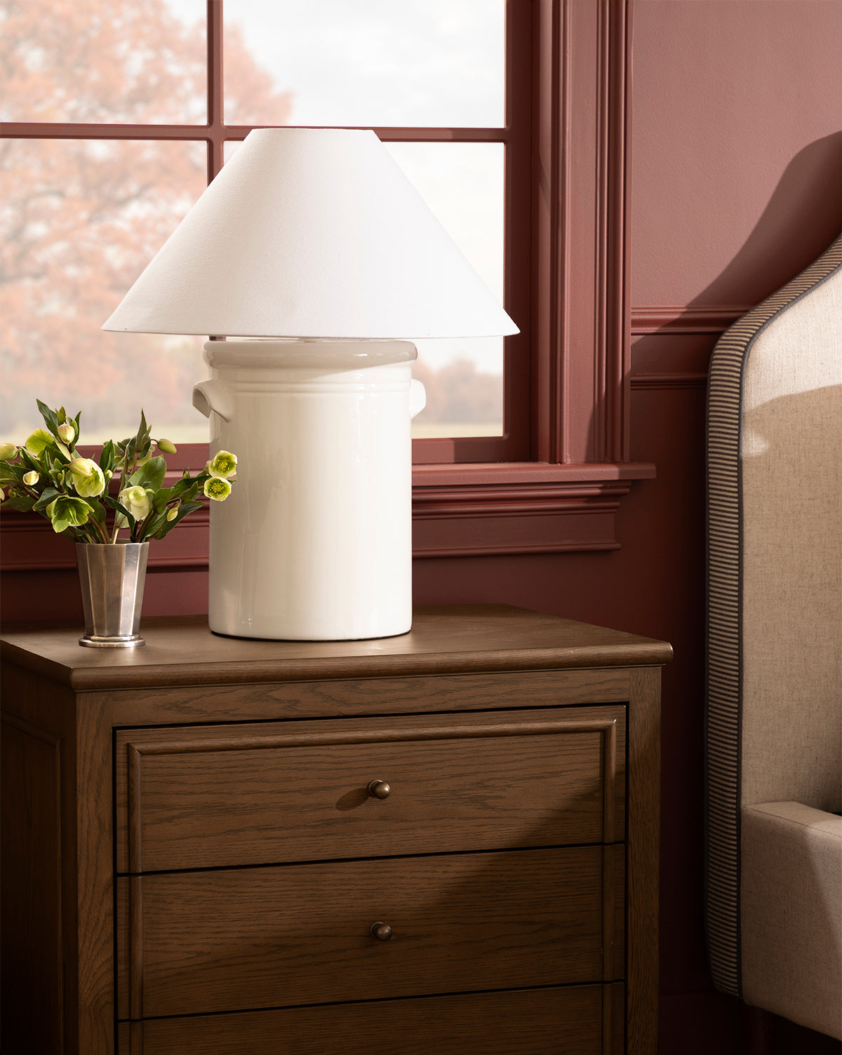 The Arles Table Lamp elevates your wooden nightstand with its elegant ceramic base and conical shade, complementing a small vase of flowers, an upholstered bed, and a window with reddish-brown trim, by Vendixs