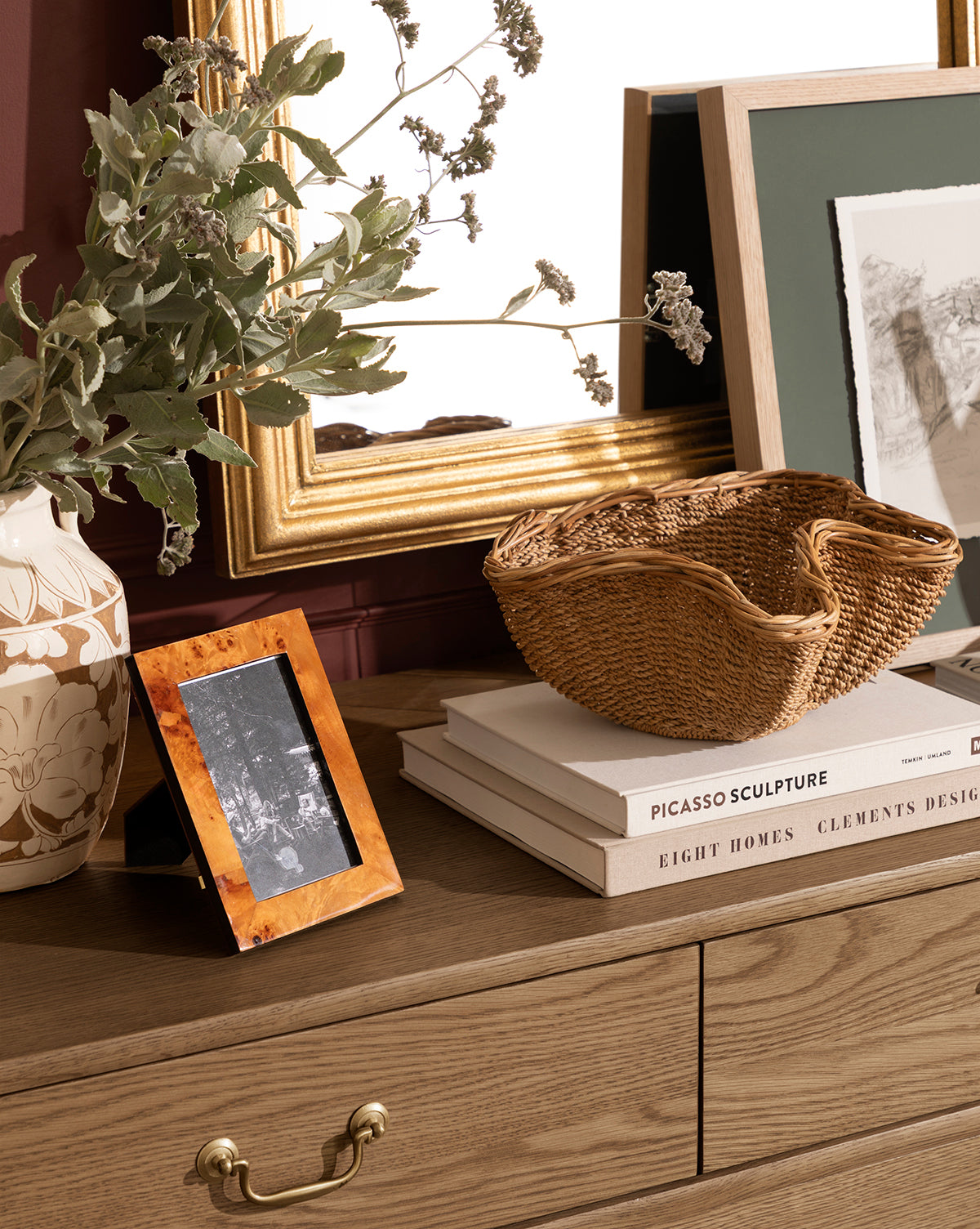 A wooden dresser with a gold-framed mirror, entryway decor like a ceramic vase with greenery, the Goulding Woven Bowl, a framed photo, and stacked books including one titled PICASSO SCULPTURE, by Vendixs