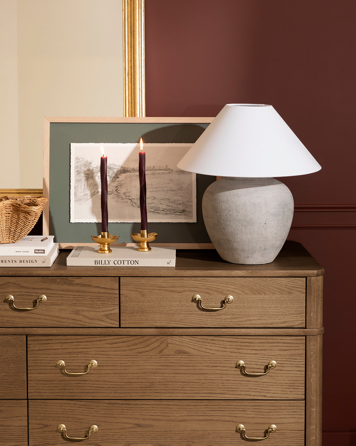 The Cove in Charcoal artwork in a light oak frame pairs with a wooden dresser featuring gold handles, a table lamp, two lit red candles in gold holders, books, and a woven basket—an inviting home decor arrangement against a burgundy wall by Vendixs