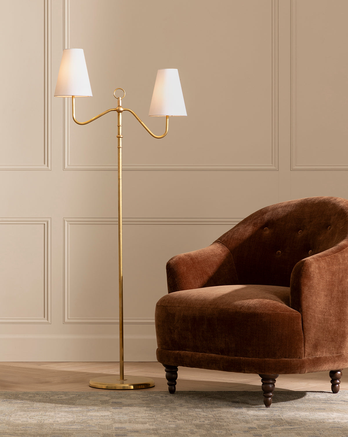 The Makerspalm Holloway Floor Lamp, in antique brass with a double-arm and two white shades, stands next to a brown velvet armchair with wooden legs on a patterned rug beside a beige paneled wall, Vendixs