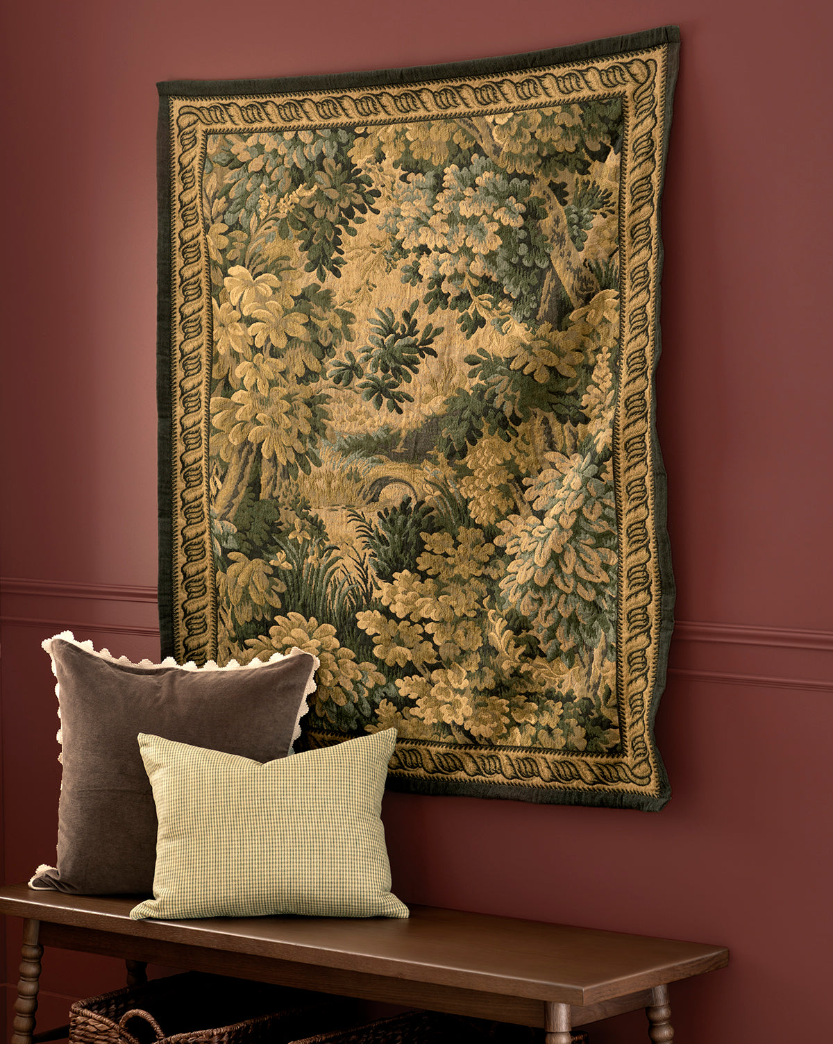 The Hortensia Wall Tapestry, featuring a leafy forest scene, hangs on a red wall above a wooden bench with two throw pillows: one brown with a scalloped edge and one light green with a checkered pattern, by Vendixs