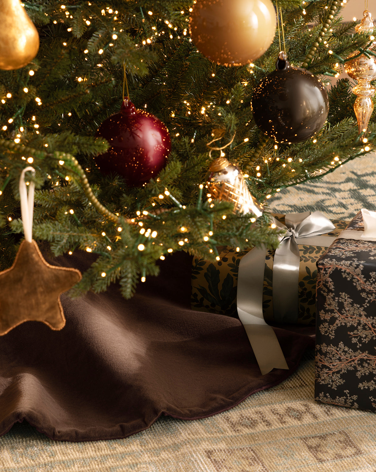 Close-up of a Christmas tree adorned with gold, red, and brown ornaments and glowing lights, with ribbon-wrapped presents on a luxurious Velvet Brown Tree Skirt resting upon a patterned rug, by Vendixs