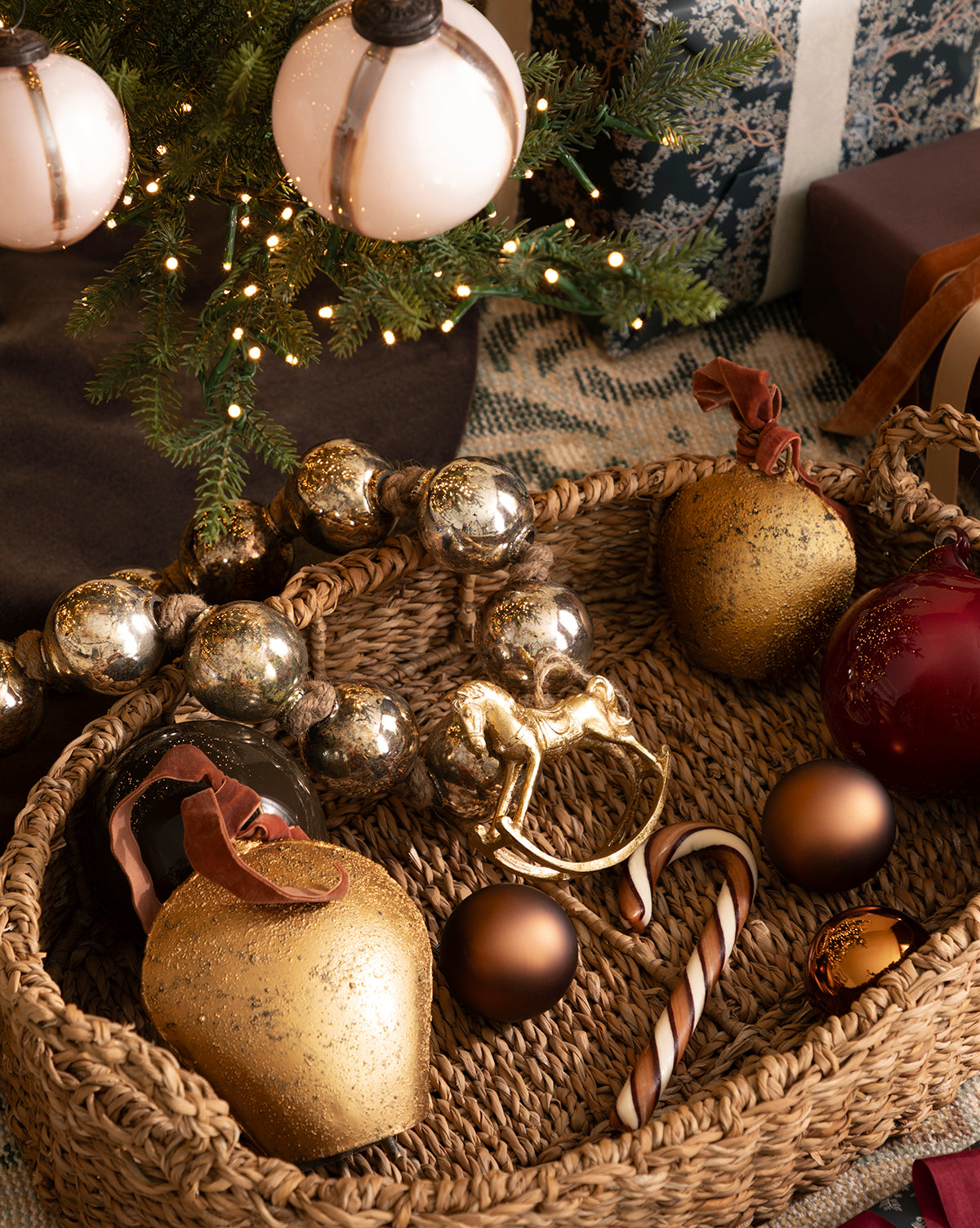 A woven basket with gold, silver, and red holiday décor sits under a decorated Christmas tree, accented by the Antique Gold Metal Bell with Velvet Ribbon for extra festive charm, by Vendixs Wrapped gifts are placed nearby.