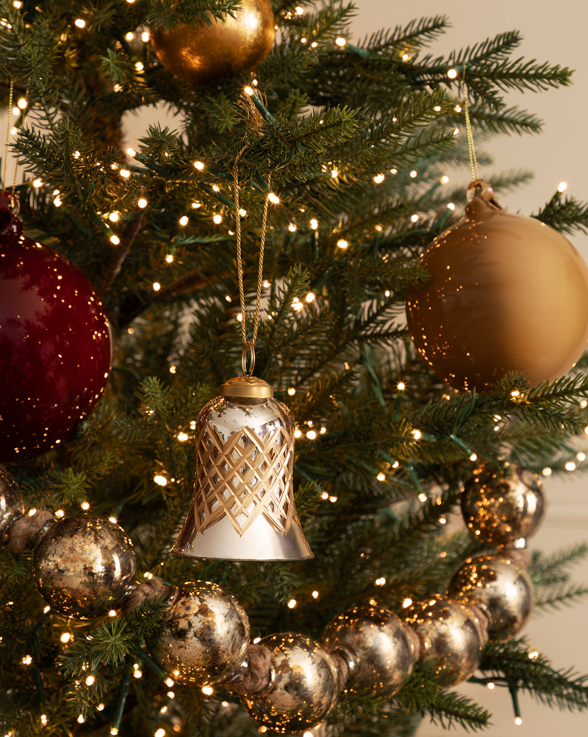 A close-up of a decorated Christmas tree featuring gold and red ornaments, a Hand-Painted Mercury Glass Bell Ornament, warm white string lights, and a garland of shiny silver beads creates festive holiday décor, by Vendixs