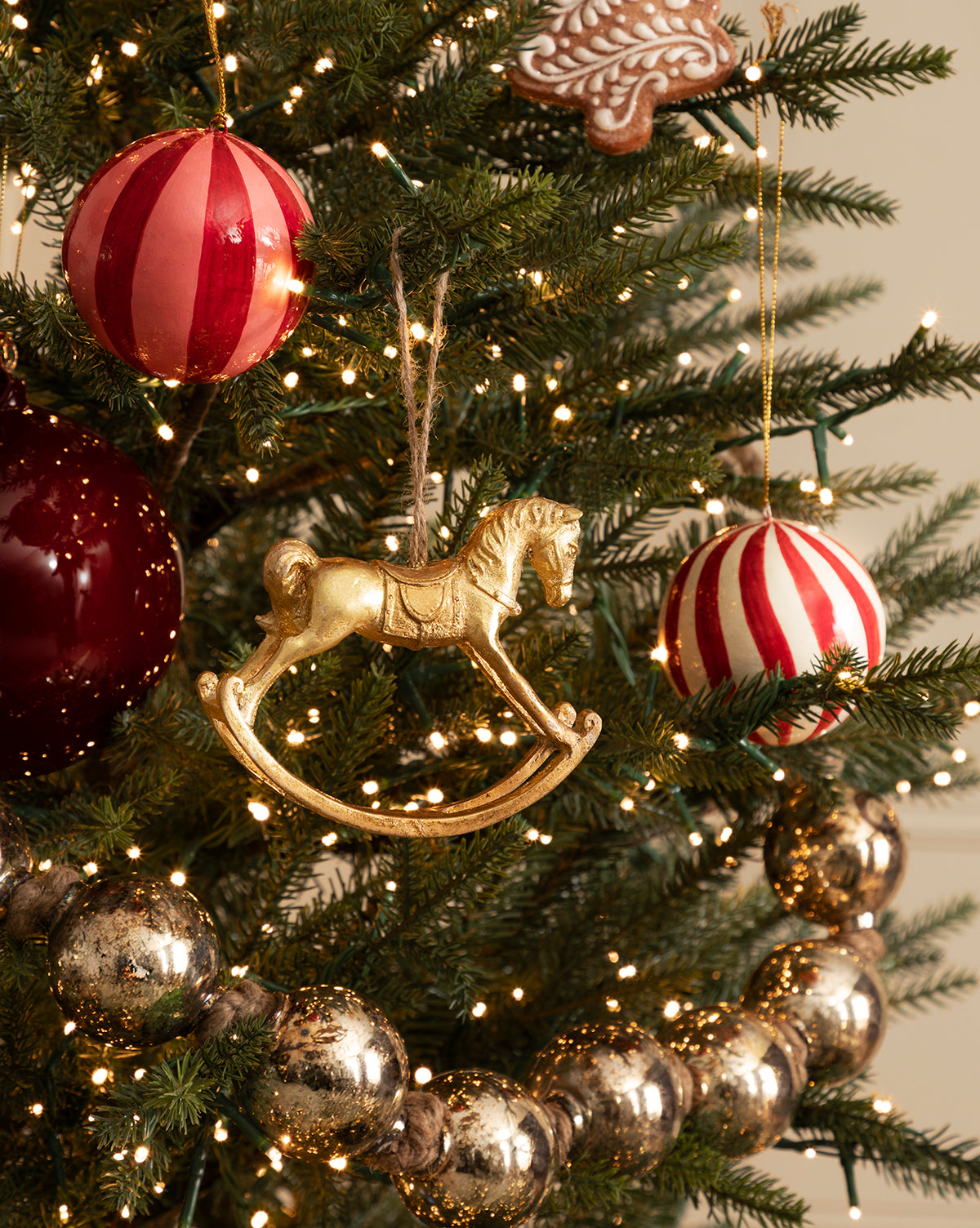 A Christmas tree decorated with 3" Hand-Painted Paper Mache Baubles with Tassel (Set of 3), gold rocking horse and gingerbread cookie ornaments, mercury glass bead garland, and warm white string lights, by Vendixs