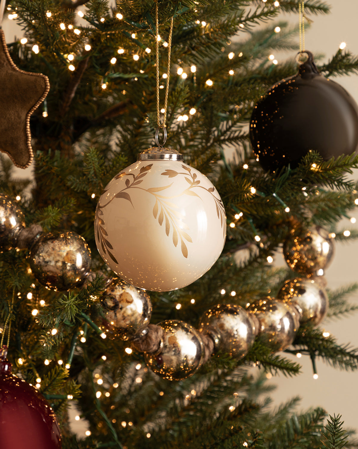 A close-up of a decorated Christmas tree with the 4" Hand-Painted Vine Glass Bauble, beaded ornaments, metallic garland, Fish with Sequins Ornaments, a black ornament, and glowing string lights for festive holiday décor, by Vendixs