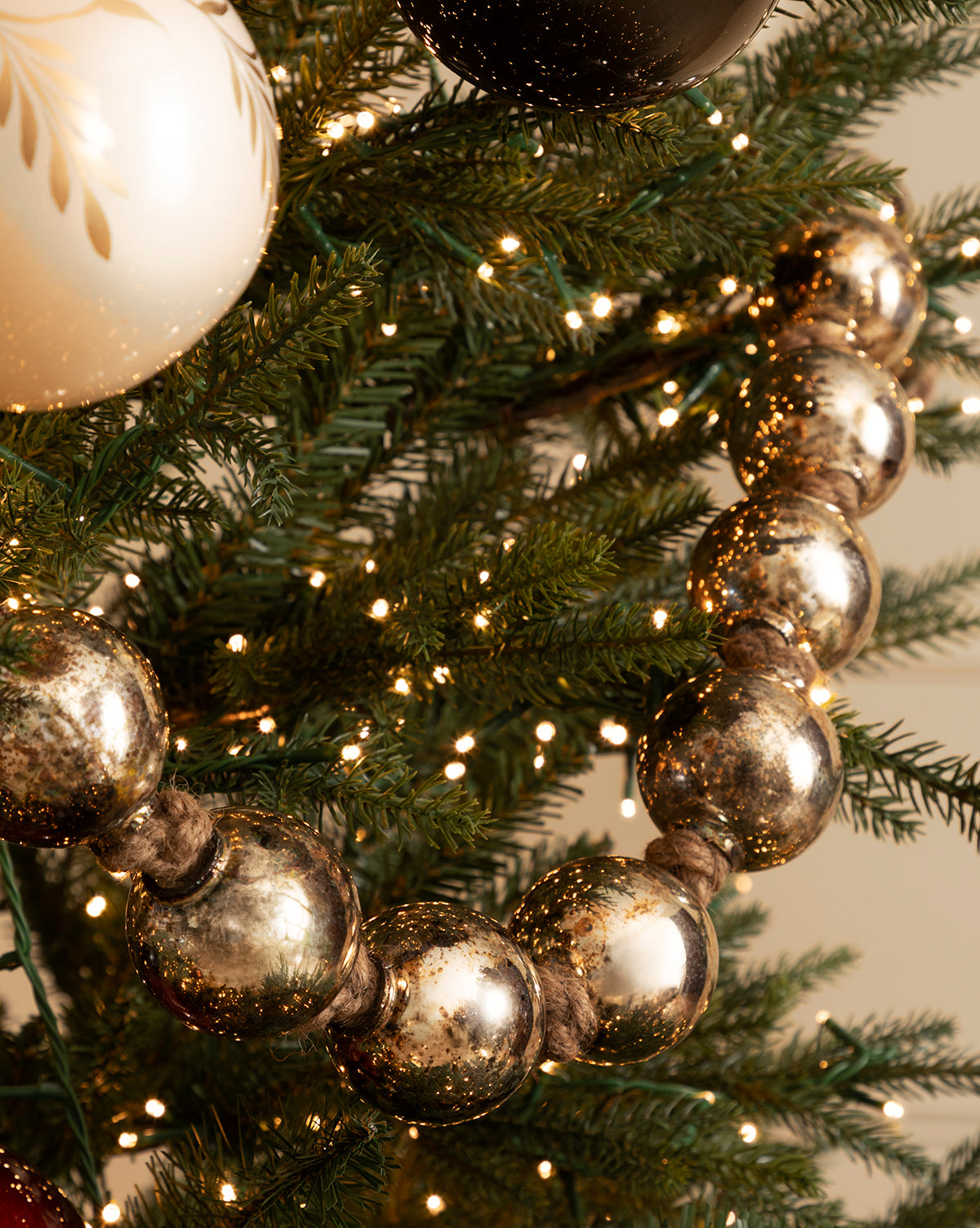 A close-up of a Christmas tree adorned with string lights, vintage holiday decor, and the 4.4' Aged Silver Glass Garland featuring large reflective beads, plus a cream-colored ornament with gold accents, by McGee & Co.