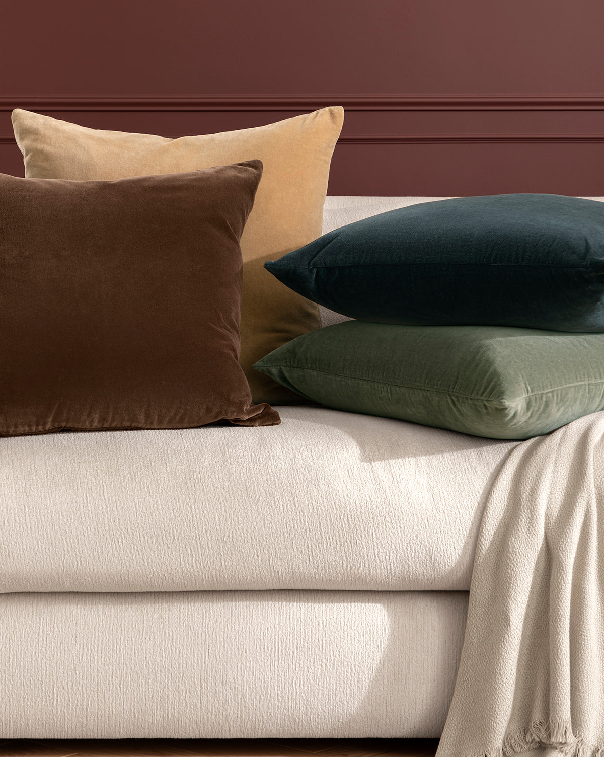 Four Bellevue Velvet Pillow Covers in brown, beige, dark green, and olive green add elegance to a light upholstered sofa with a cream blanket, set against a dark reddish-brown wall, by Vendixs