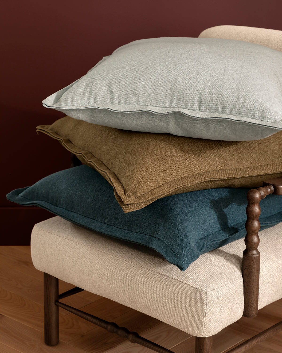 Three large Liam Double Flange Pillow Covers in light gray, mustard yellow, and deep teal are stacked on a beige upholstered bench with a wooden leg and spindle arm, set against a burgundy wall and wood floor, by Vendixs