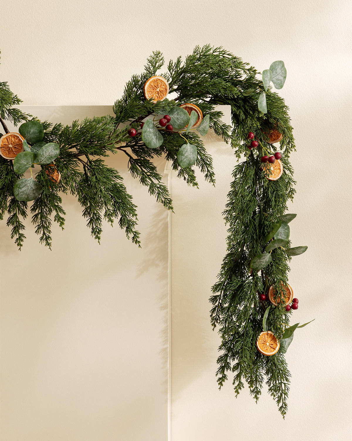 The 6' Orange & Eucalyptus Faux Garland, crafted with evergreen branches, eucalyptus leaves, red berries, and dried orange slices, adds festive flair to any light-colored wall or mantel, by Vendixs