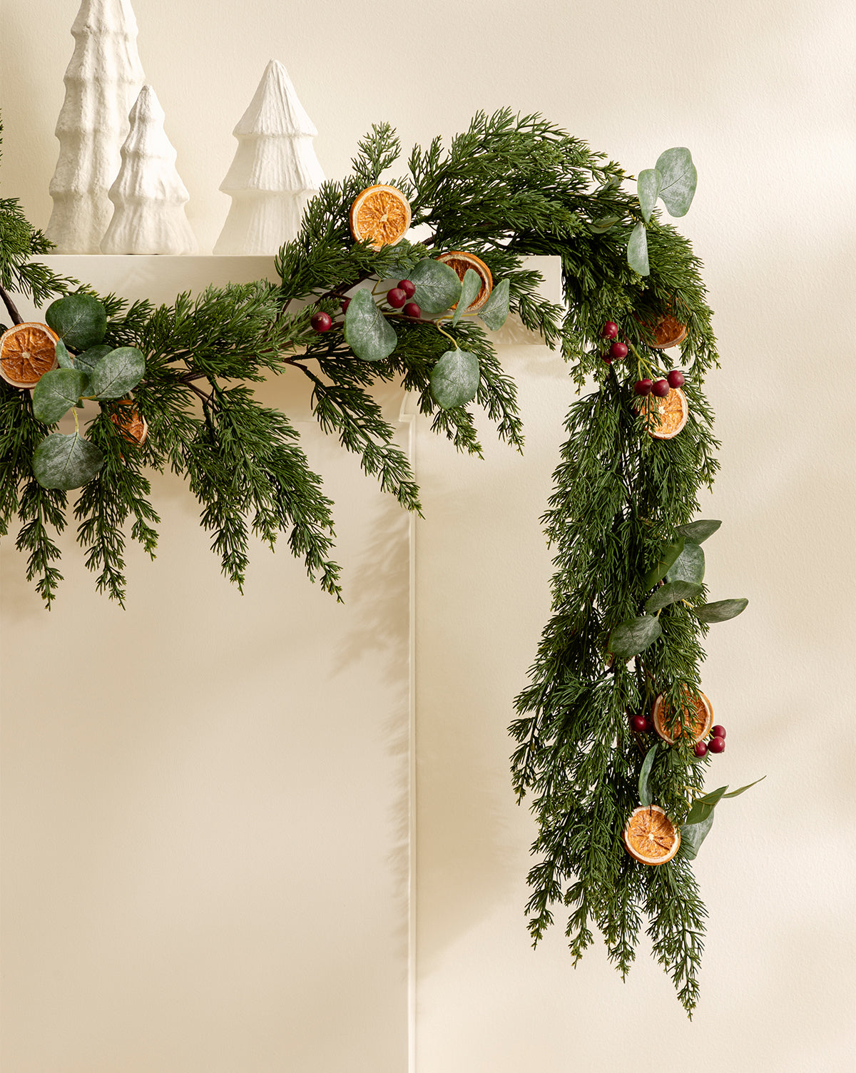 The 6' Orange & Eucalyptus Faux Garland, featuring pine branches, red berries, and dried orange slices, is draped over a white mantel and paired with white ceramic Christmas trees for festive seasonal décor, by Vendixs