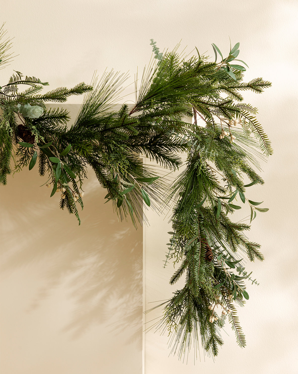 A close-up of the 6' Pinecone & Berries Faux Garland, featuring lush evergreen branches, pinecones, and red berries, draped diagonally on a light beige wall—ideal for festive holiday décor, by Vendixs
