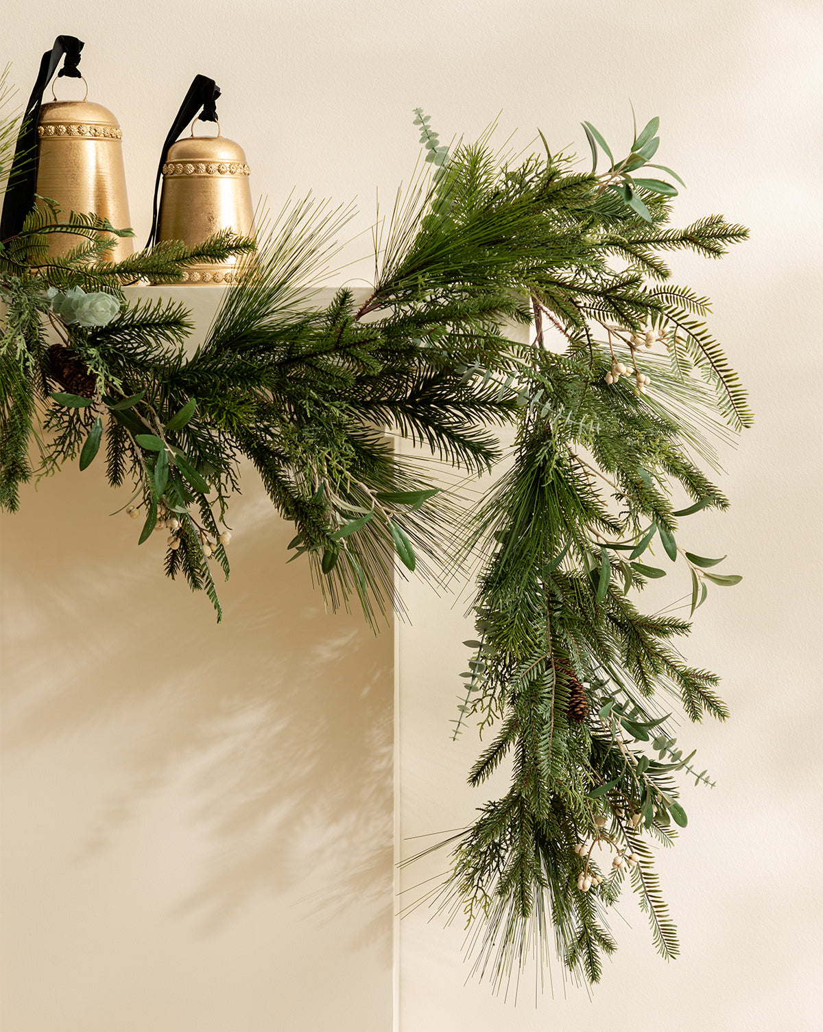 The 6' Pinecone & Berries Faux Garland drapes over a light mantel, accented with two large gold bells and black ribbons for charming holiday décor, by McGee & Co.
