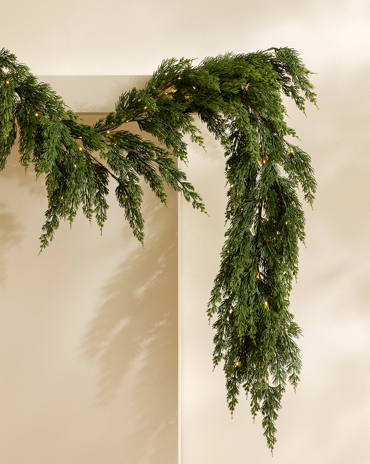 The 6' Cedar Pre-Lit Faux Garland drapes over a light beige doorway, adding festive and natural holiday decor with its green, leafy look against the neutral wall, by Vendixs
