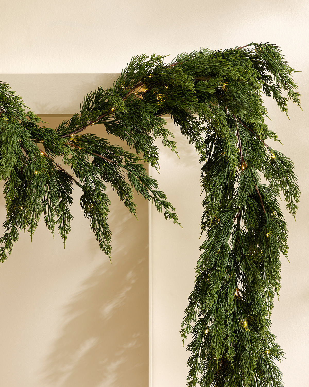 The 6' Cedar Pre-Lit Faux Garland, adorned with small warm white lights, drapes beautifully over a doorway against a light wall—ideal for festive holiday home decor, by Vendixs