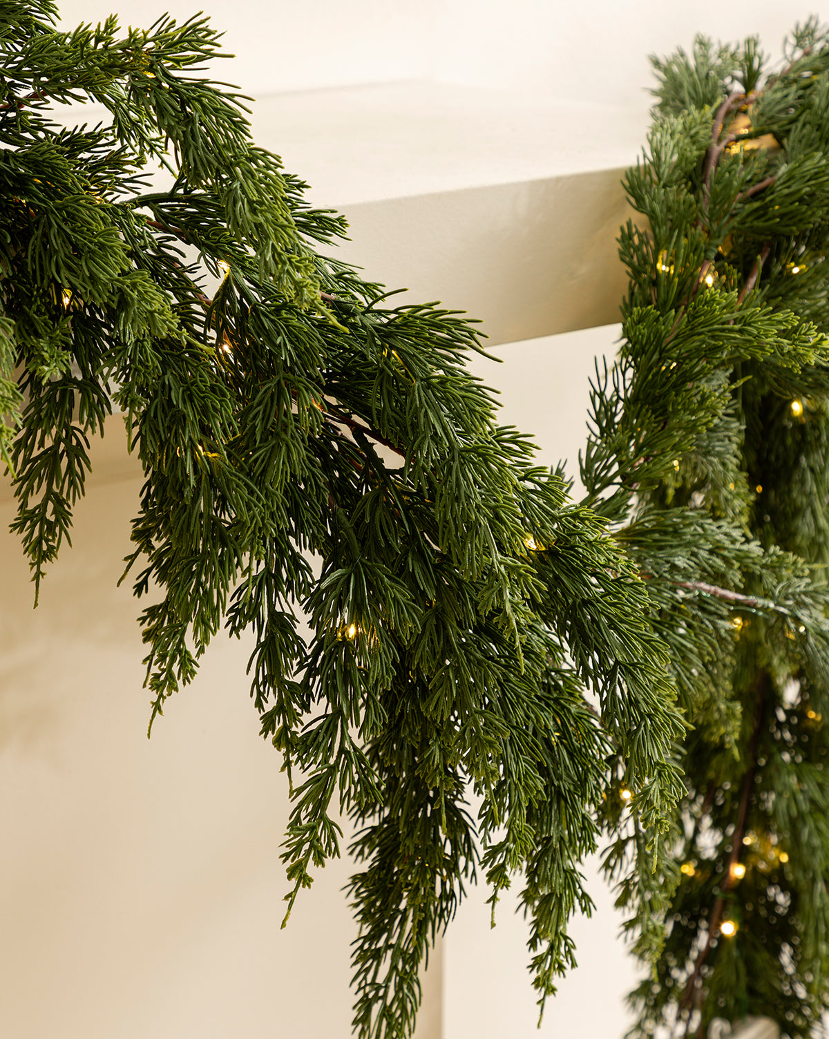 A close-up of the 6' Cedar Pre-Lit Faux Garland, featuring small warm white lights draped over a white mantel, brings festive charm to your holiday home décor with its lush, realistic design, by Vendixs