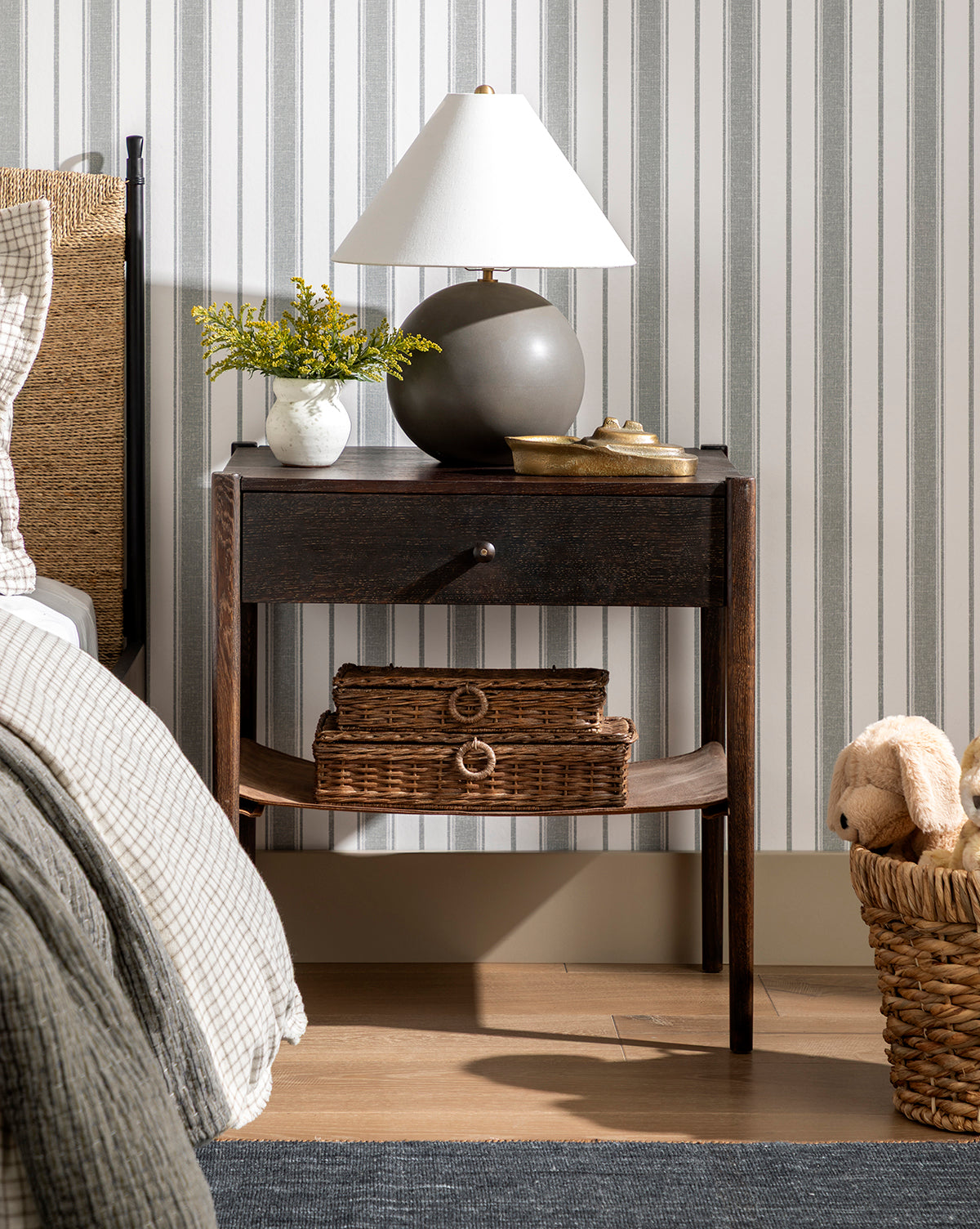 A dark wooden nightstand beside a bed holds a Tess Ceramic Table Lamp, a small vase with yellow flowers, and a dish. Two wicker baskets are on the lower shelf, and a basket with a stuffed animal sits nearby on the floor, by Vendixs