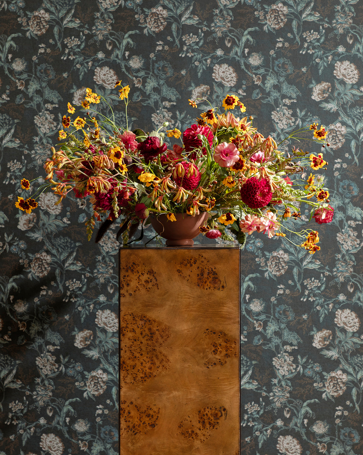 A large, colorful bouquet of wildflowers and peonies in a brown vase sits atop the Eamon Pedestal, standing before dark floral wallpaper with flowers spilling out in all directions, creating a vibrant, dramatic display, by Vendixs