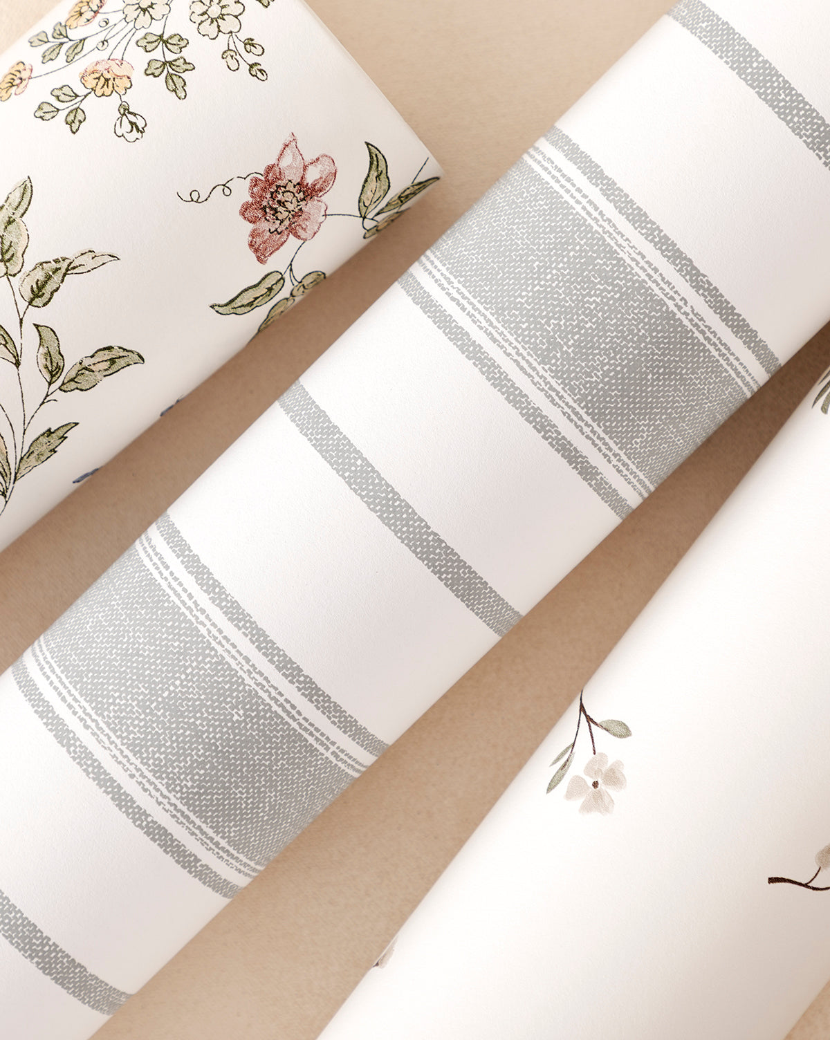 Three rolls of wallpaper are displayed: one with a floral pattern, the Hawes Stripe Wallpaper with classic gray and white vertical stripes, and one with a small flower design, all set on a light beige surface by Vendixs