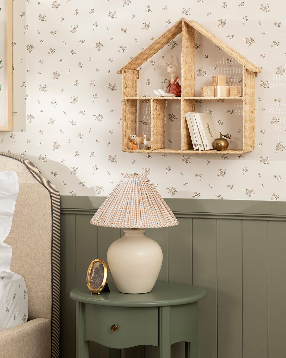 A cozy bedroom corner features a green nightstand with a lamp and framed photo. Above, the house-shaped Maison Shelf in natural finish showcases decor and books against floral wallpaper, by Vendixs