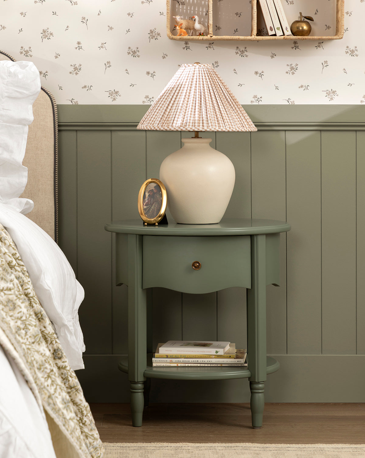 A green Liberty Nightstand holds a beige lamp with a striped shade, a round photo frame, and stacked books. GREENGUARD Gold Certified, this table sits beside a bed against a floral and paneled wall, by Vendixs