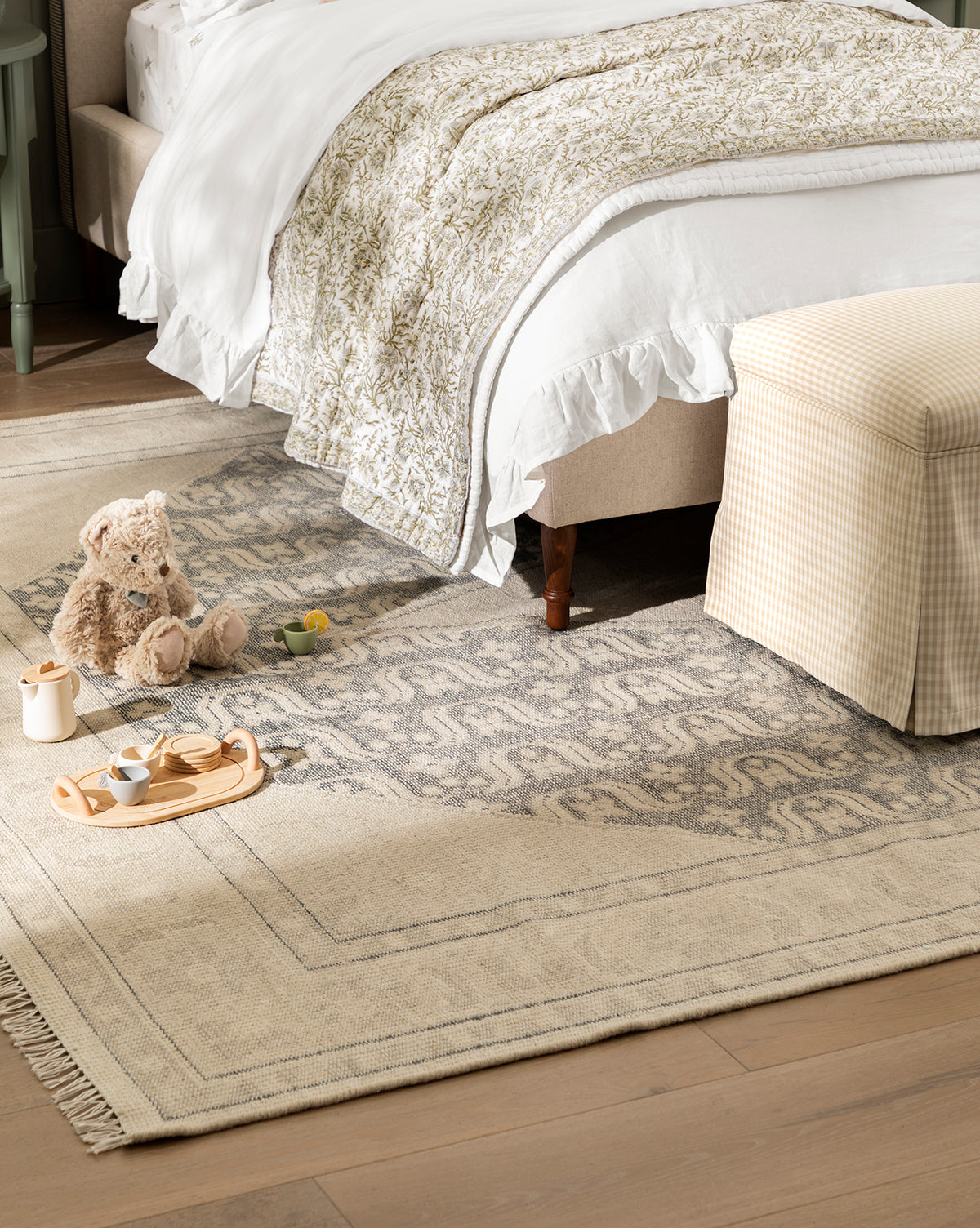 A cozy bedroom features a bed with white and patterned bedding, a pale plaid ottoman, and a plush teddy bear beside a wooden tea set atop the artisan-inspired Elin Hand-Knotted Wool Rug, by Vendixs