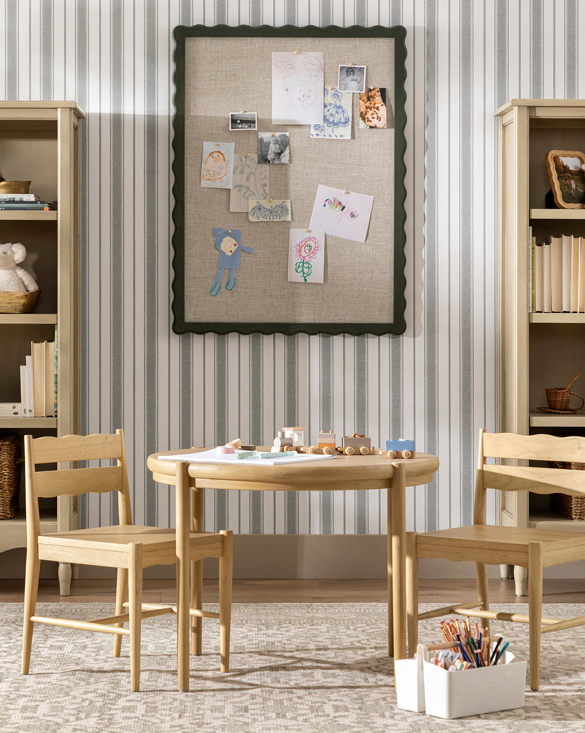 A children’s playroom features a round white oak Darcy Kids Table with two chairs, art supplies, and toy cars. Behind it, a bulletin board displays drawings and photos, flanked by bookshelves on striped wallpaper, by Vendixs
