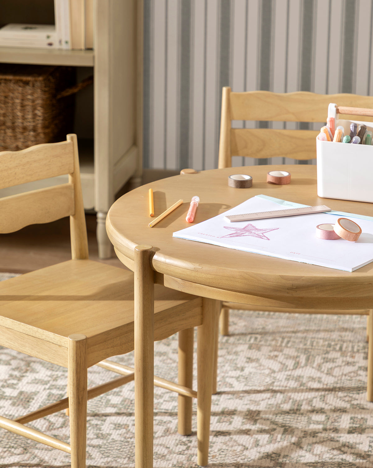 The Darcy Kids Table, paired with two chairs, displays art supplies, crayons, and markers. A star drawing and a white supply container rest on the table. Striped wallpaper and a woven basket are seen in the background, by Vendixs