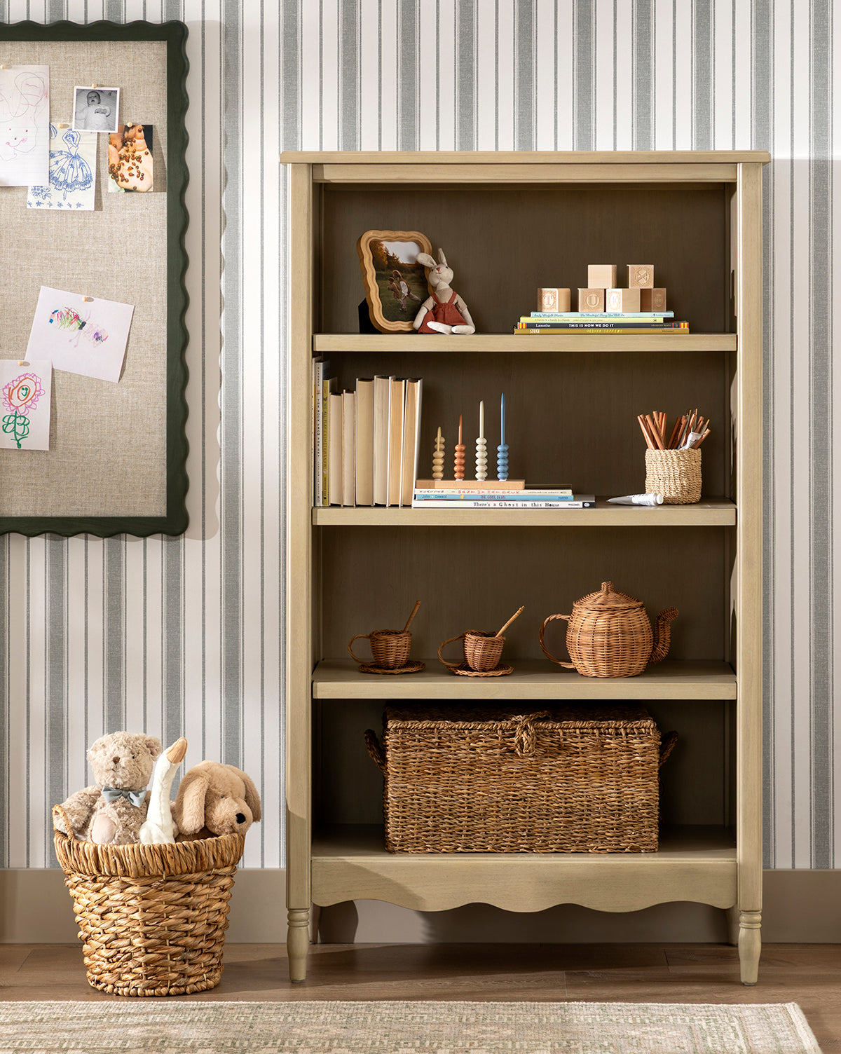 A bookshelf with books, decor, and handwoven baskets stands against striped wallpaper. A Water Hyacinth Basket with Handles (Set of 2) filled with stuffed animals sits nearby; childrens drawings are pinned on a bulletin board to the left, by Vendixs