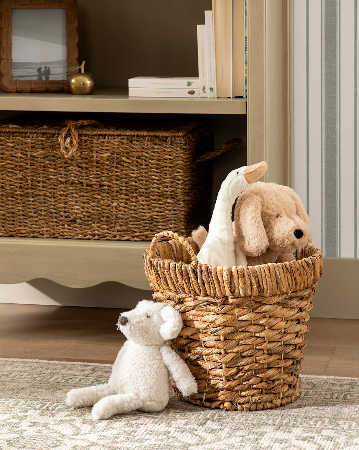 A decorative storage basket with stuffed animals—a dog and a duck—sits on a rug by a white teddy bear. Nearby, Water Hyacinth Baskets with Handles (Set of 2) and books add charm to the shelf with framed decor, by Vendixs
