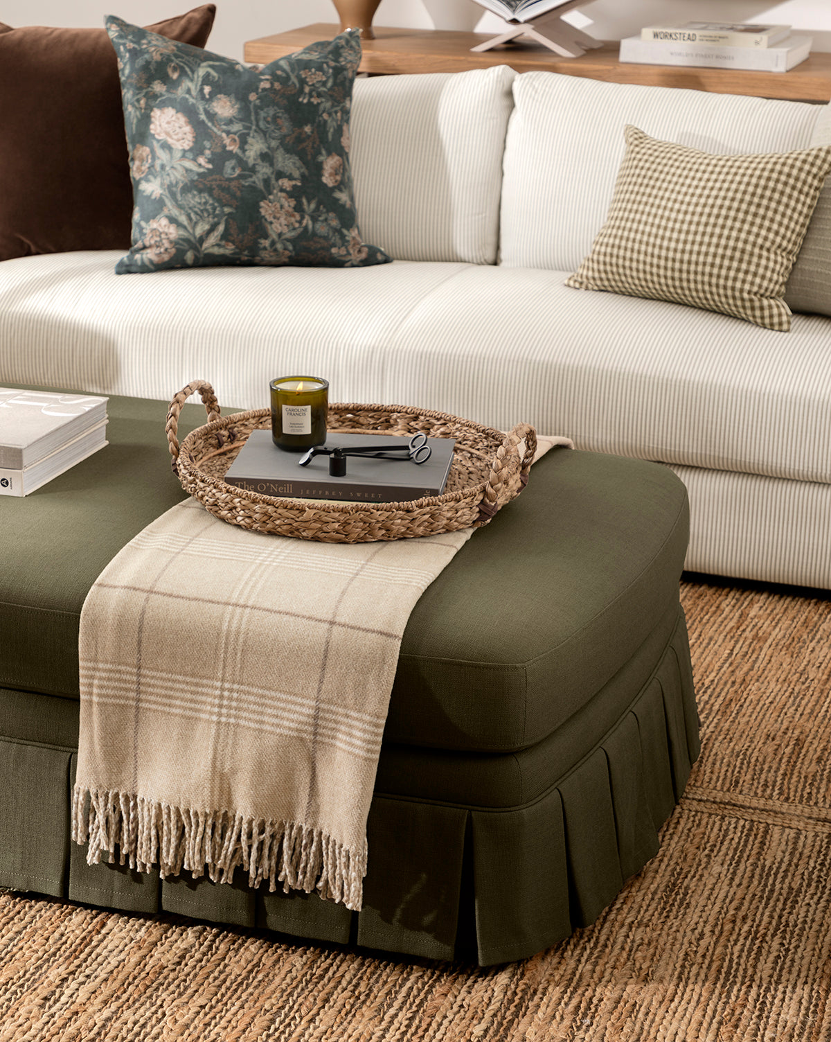 A cozy living room has a white sofa with patterned pillows, a green ottoman with a beige plaid throw, and the Salter Woven Tray displaying a candle, book, and glasses. A textured rug completes the inviting space, by Vendixs