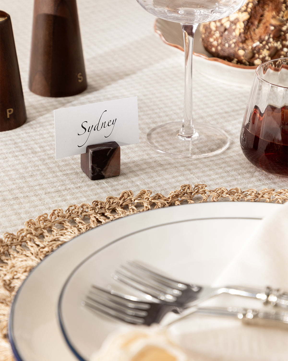 A close-up of an elegant holiday table features Auburn Marble Place Card Holders (Set of 4) with a card reading Sydney, forks on a white plate atop a woven placemat, and wine glasses nearby, by Vendixs