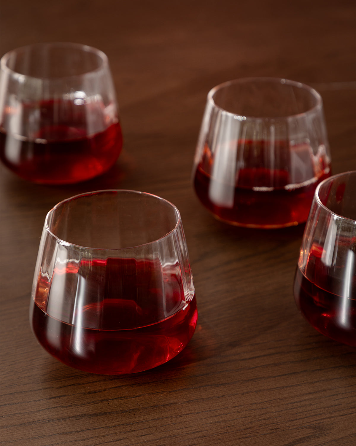 A set of four Langford Tumblers, filled with red liquid, rests on a wooden surface—their reflections shimmering and highlighting the elegance of this glassware set by Vendixs