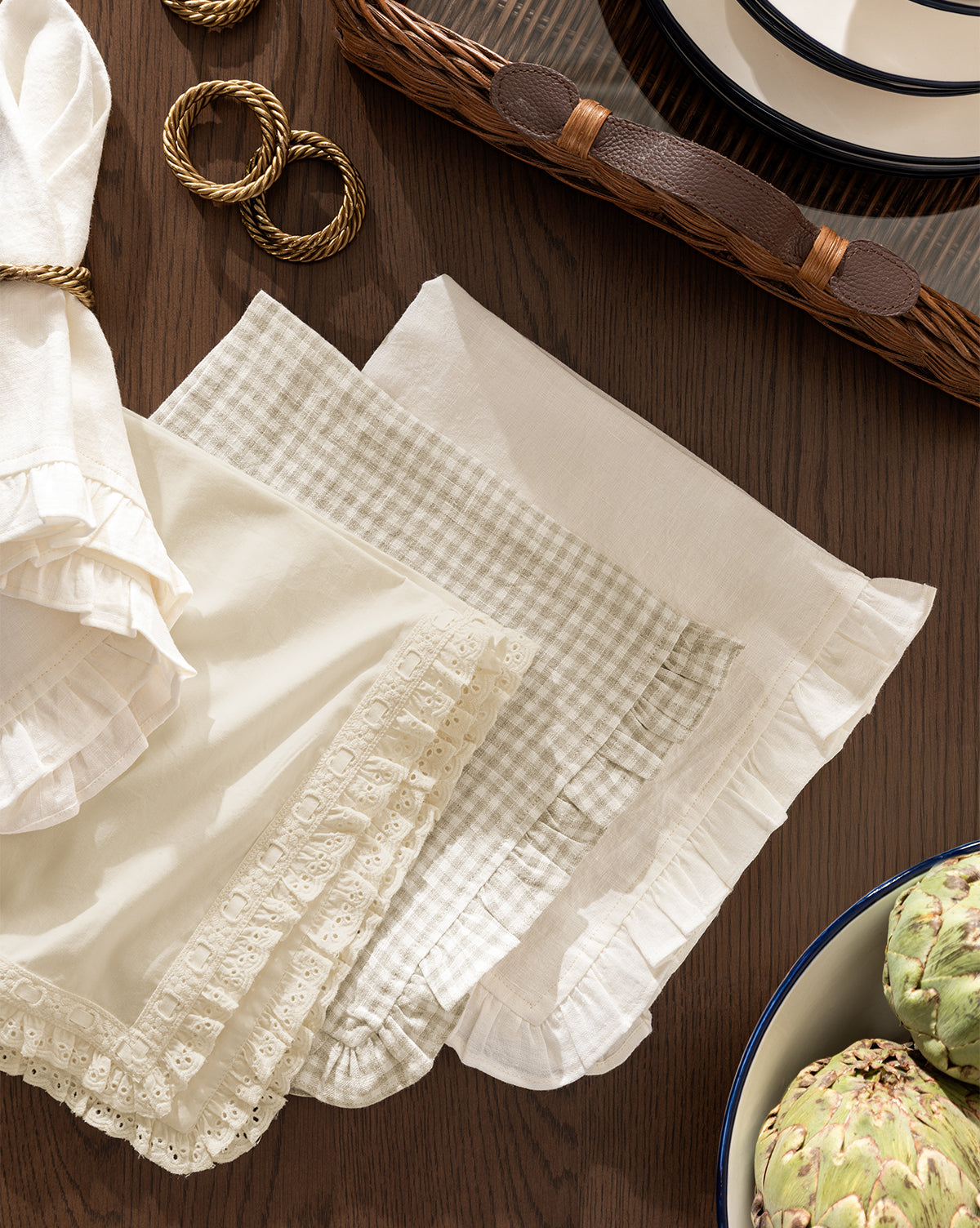 The Lace Trim Napkins (Set of 4), featuring beige and white vintage-inspired fabric with ruffled lace edges, are folded on a wooden table, paired with gold napkin rings, stacked plates in a wicker tray, and a bowl of artichokes by Vendixs