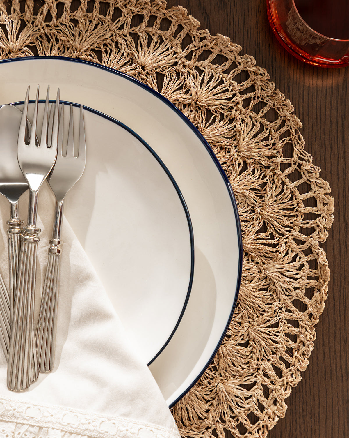 Overhead view of two stacked white plates with dark rims on the Hermione Placemat, topped with a folded napkin and three silver forks, next to a glass containing a reddish drink by Vendixs