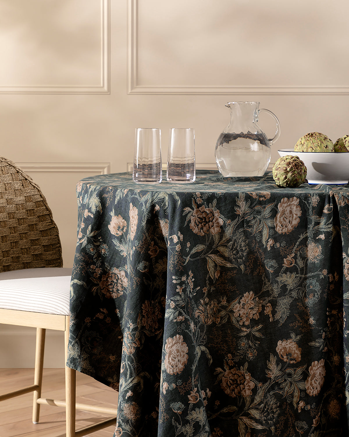 A round table draped with a dark Hestercombe Tablecloth holds a clear glass pitcher, two empty glasses, and a white bowl of artichokes. A woven chair with a white cushion sits beside the table against a beige wall with molding, by Vendixs