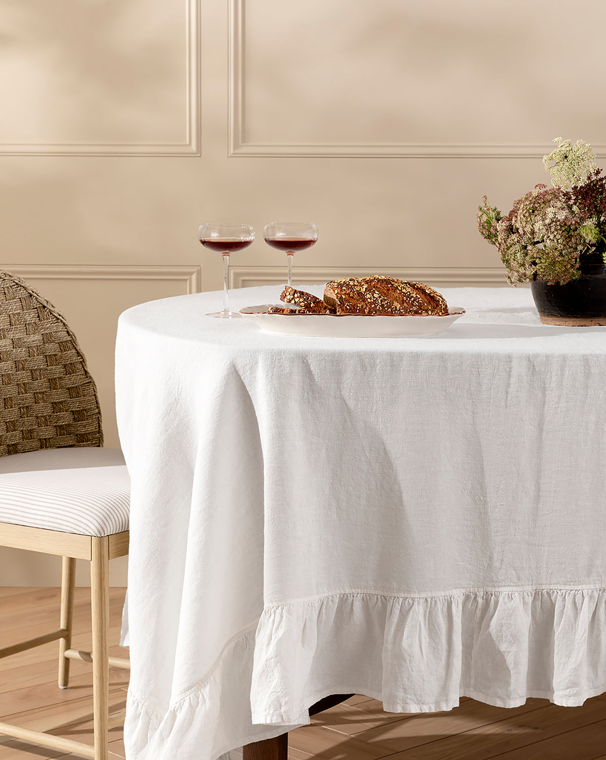 A round table draped with the Catania Linen Ruffle Tablecloth is set with two glasses of red wine, a loaf of bread on a cutting board, and a vase of flowers. A cushioned chair with a woven back sits nearby, by Vendixs