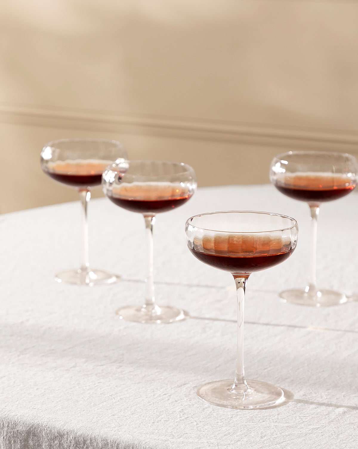 A set of four Langford Coupe glasses, each filled with a dark brown liquid, sits on a textured white tablecloth against a soft beige background—showcasing the elegant Langford Coupe (Set of 4), perfect for any occasion, by Vendixs