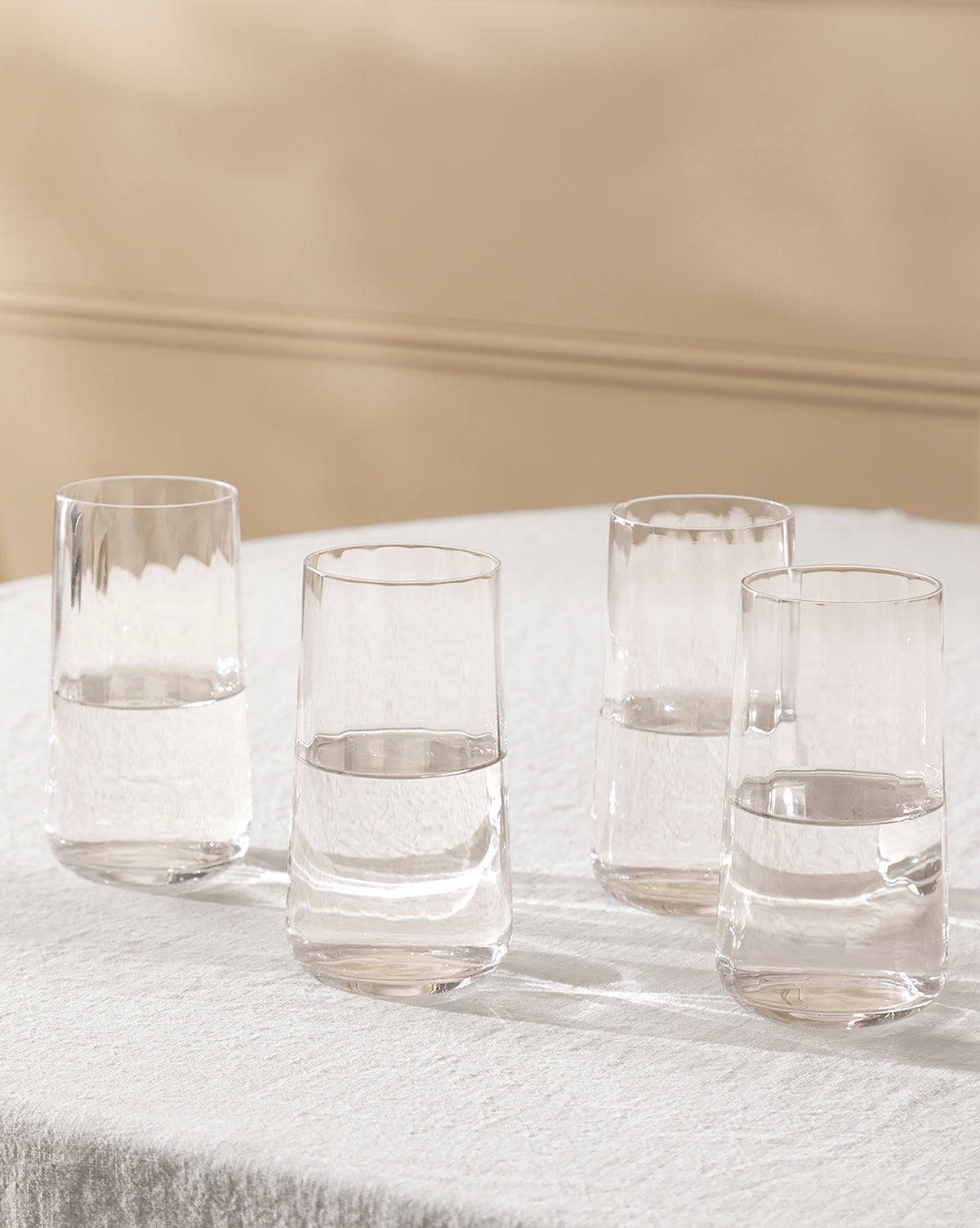 A set of four Langford Highball Glasses, each partially filled with water, is arranged on a white textured tablecloth against a soft beige background, by Vendixs