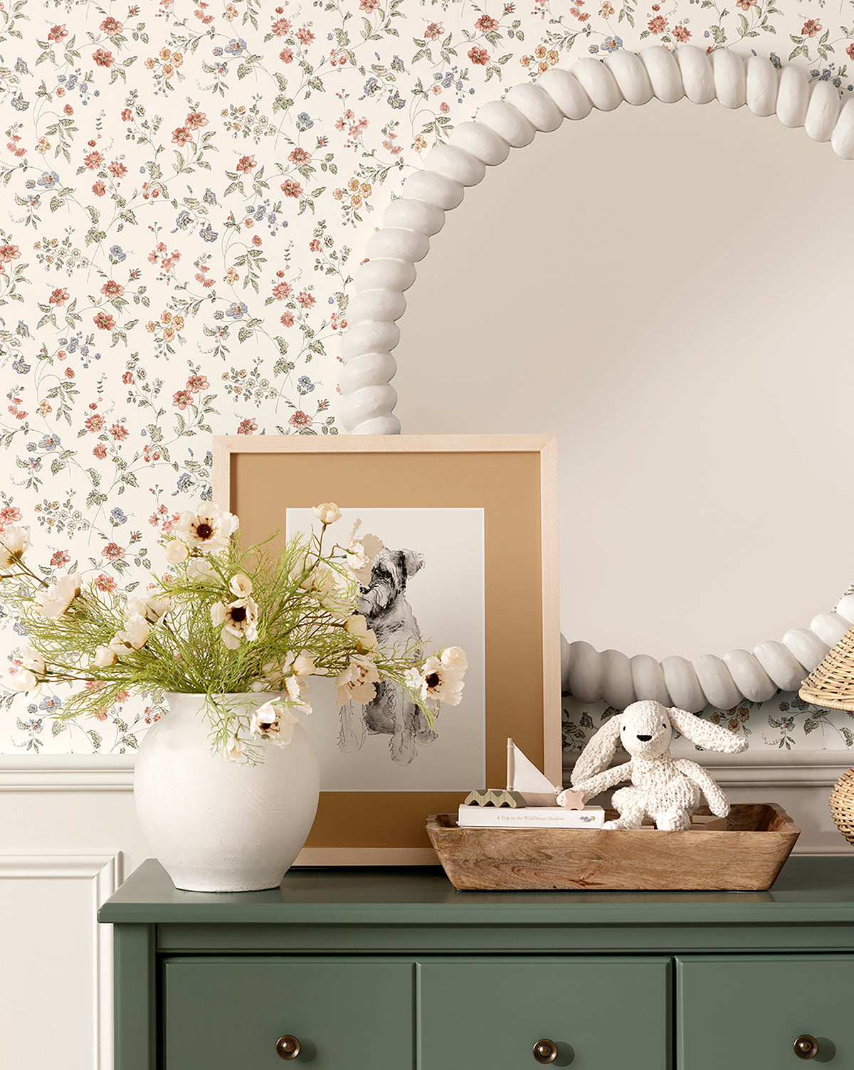 A green dresser holds a white vase of flowers, framed dog sketch, Scalloped Wood Tray with a plush bunny, and toy sailboat. Above, a large round mirror with a textured frame hangs on floral wallpaper, by Vendixs