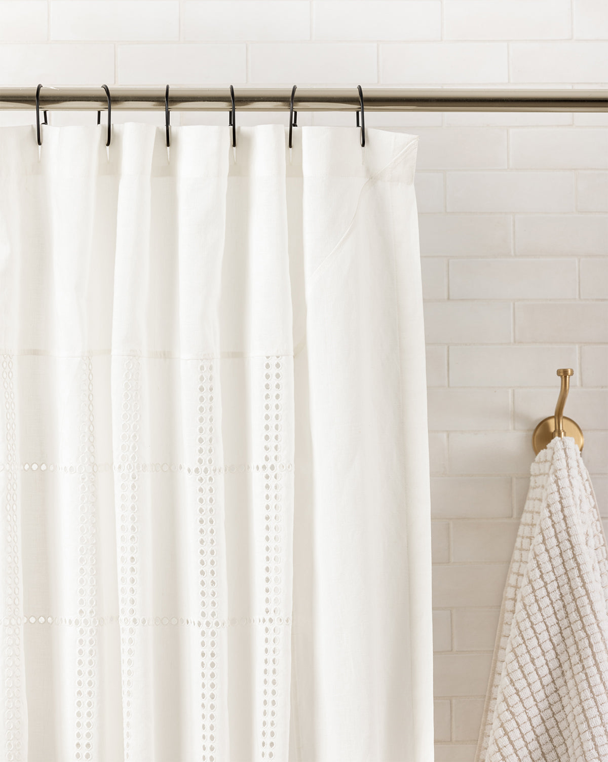 The Albany Shower Curtain, in white linen-cotton with decorative stitching, hangs from a silver rod beside a beige textured towel on a hook, set against white tiled walls for an elegant bathroom look, by Vendixs