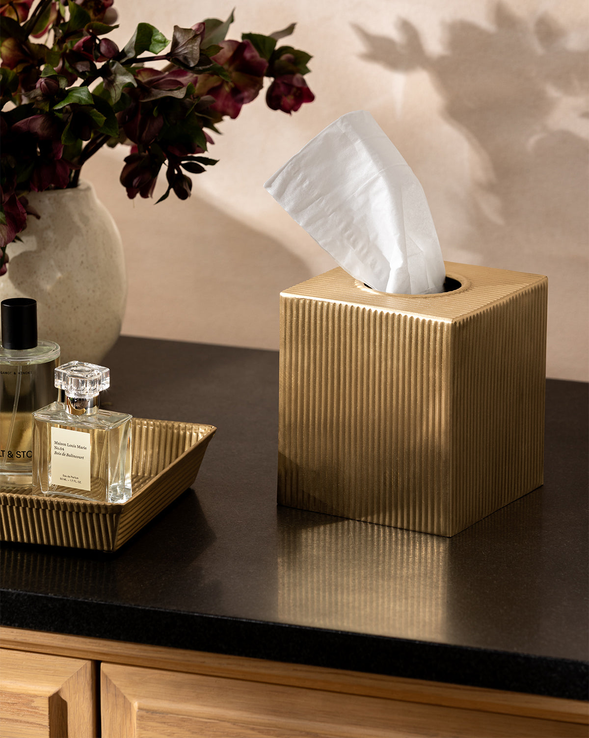 A Calvert Brass Tissue Box Cover sits on a dark countertop beside a tray of perfume bottles, with a vase of deep red flowers visible in the background, by Vendixs