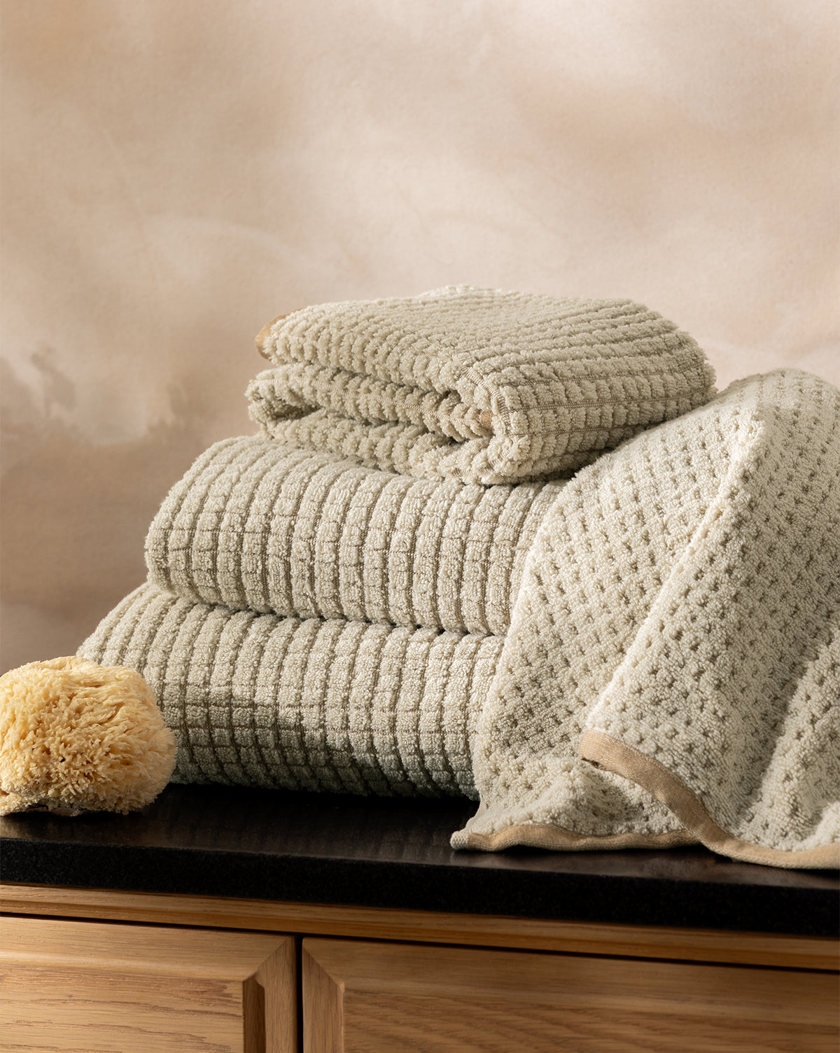 The Atrani Organic Bath Collection, featuring beige textured cotton towels folded and draped on a dark countertop beside a natural sponge, is set against light wood cabinets and a soft beige background, by Vendixs