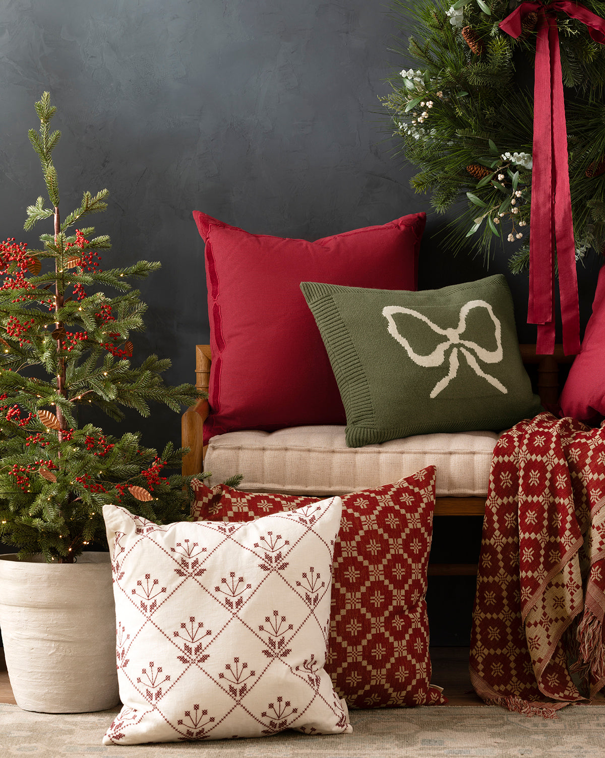 A cozy holiday scene featuring the McCallister Pillow Cover in festive red and green on a wooden bench, complemented by a mini potted Christmas tree and a wreath with a red ribbon—perfect for seasonal gatherings, by Vendixs