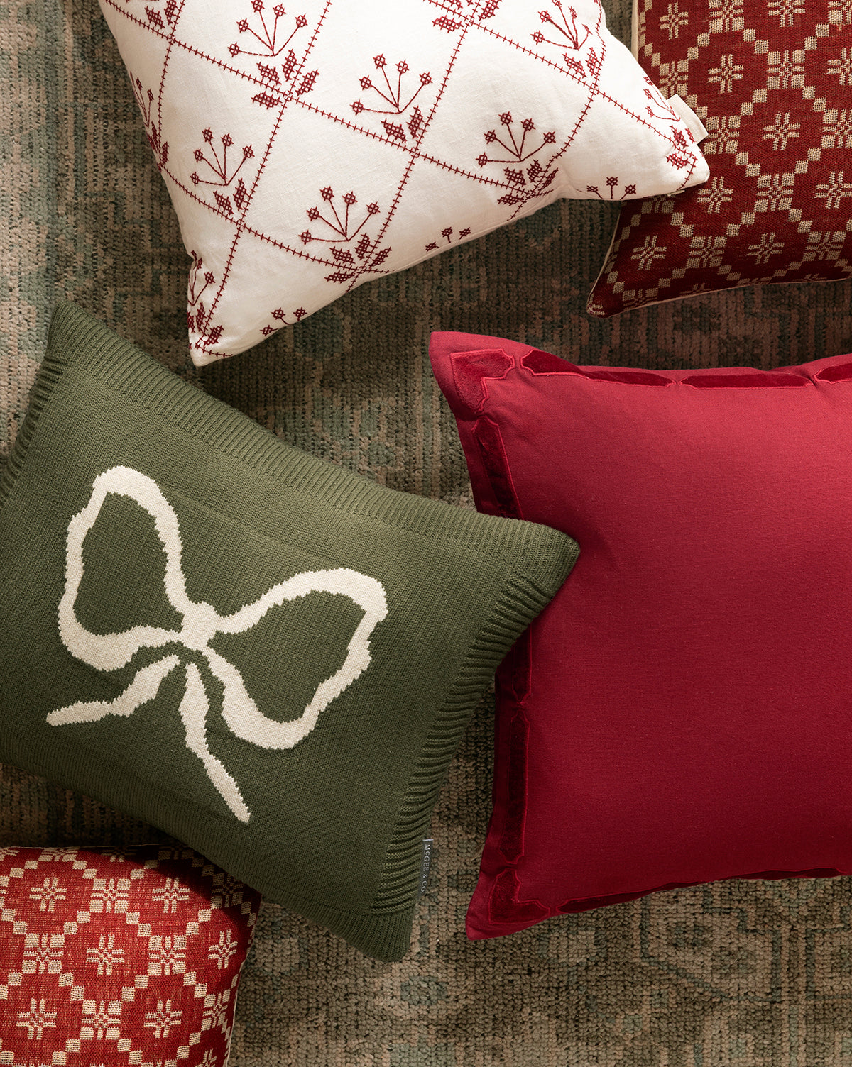 Four pillow covers are arranged on a rug, featuring festive holiday layering in red, white, and green. A green pillow with a white bow stands out among Bella Pillow Covers decorated with geometric and floral patterns, by Vendixs